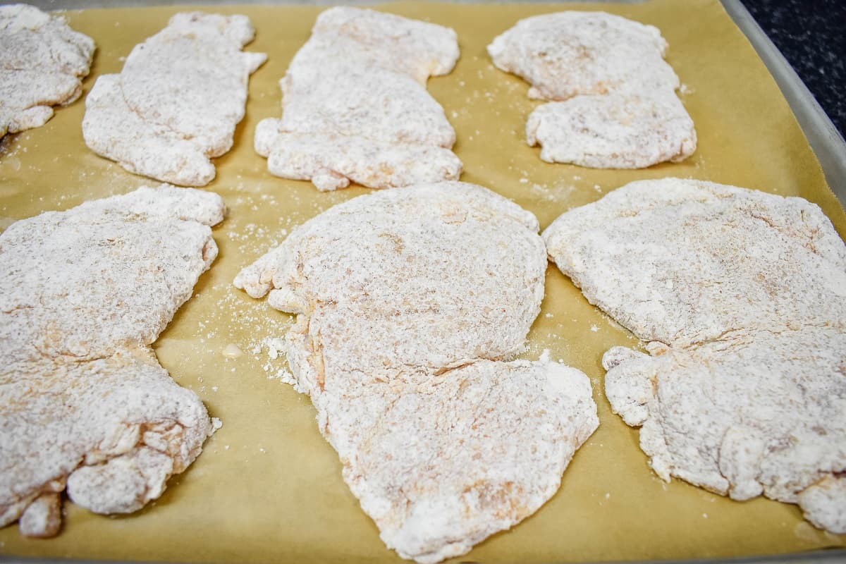 Chicken thighs coated in flour displayed on a baking sheet that's lined with brown parchment paper.