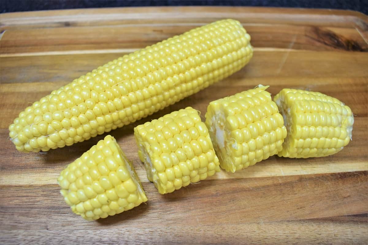 Two corn cobs, one of which as been cut into four pieces, displayed on a wood cutting board.
