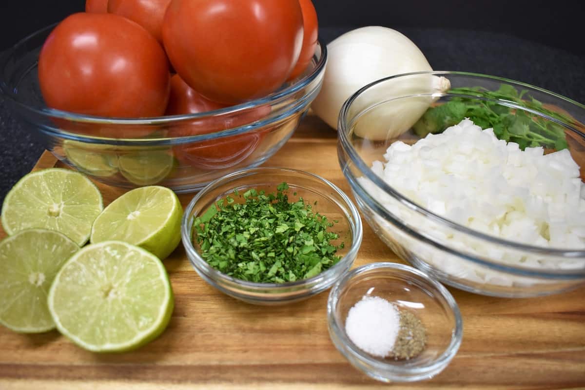 Fresh salsa ingredients, tomatoes, onions, cilantro, limes, salt and pepper on a wood cutting board.