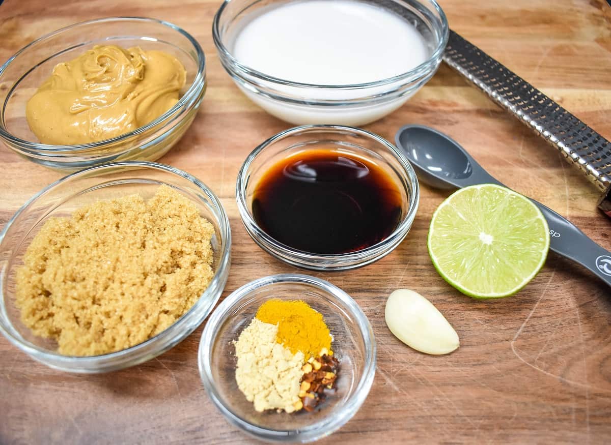 The ingredients for the peanut sauce in clear, glass bowls displayed on a wood cutting board.
