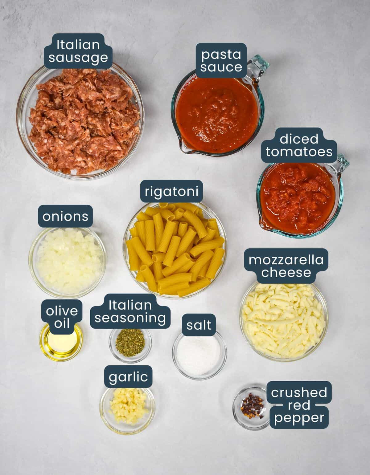 The ingredients for the recipe prepped arranged in clear bowls on a white table with each labeled with blue and white letters.