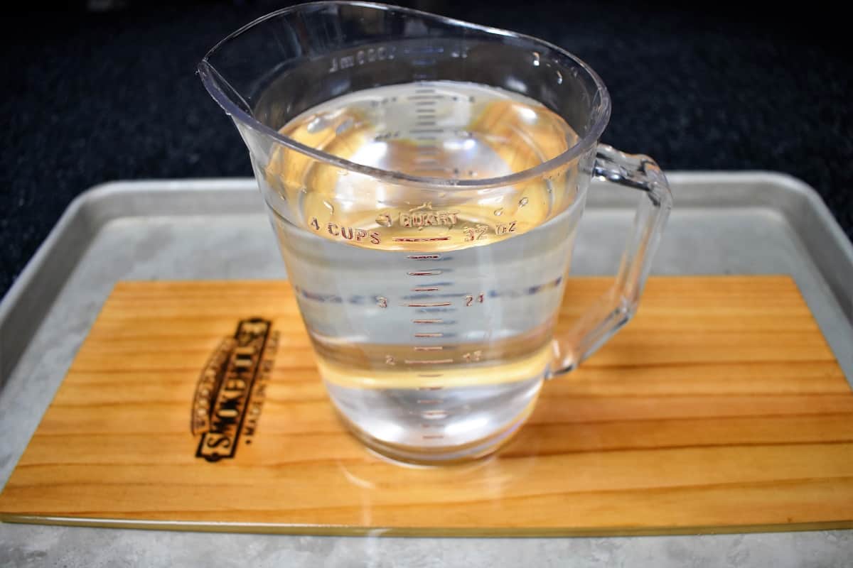 A cedar plank covered with water with a large measuring cup on top to weigh it down.