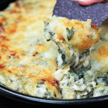 Spinach and artichoke dip served in a black skillet with some being scooped by a blue tortilla chip.