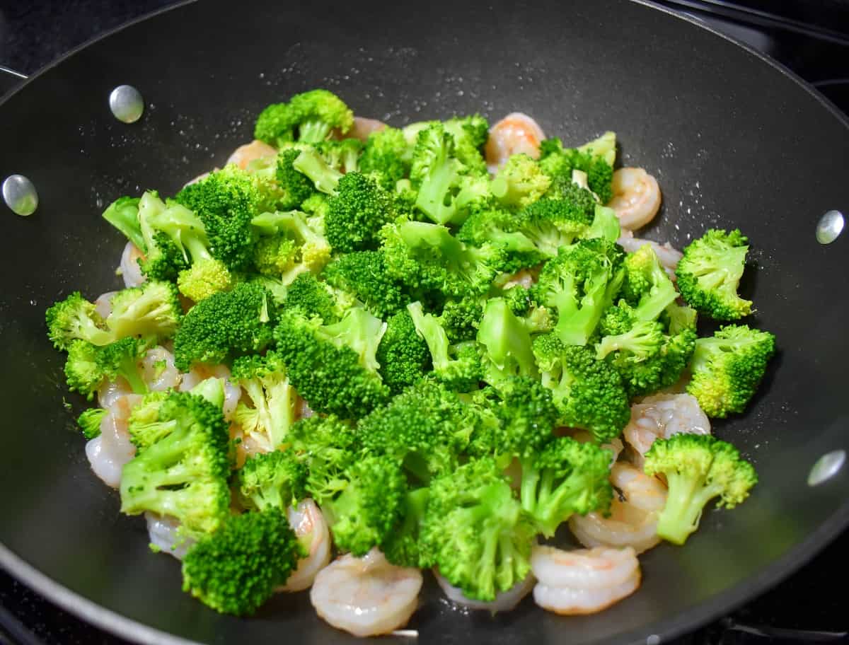 An image of broccoli florets added to shrimp in a large, black skillet.