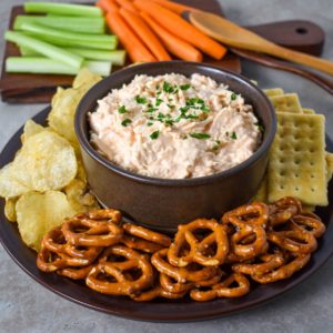 The Buffalo chicken dip served in a brown bowl served with pretzels, chips, crackers, carrots, and celery.