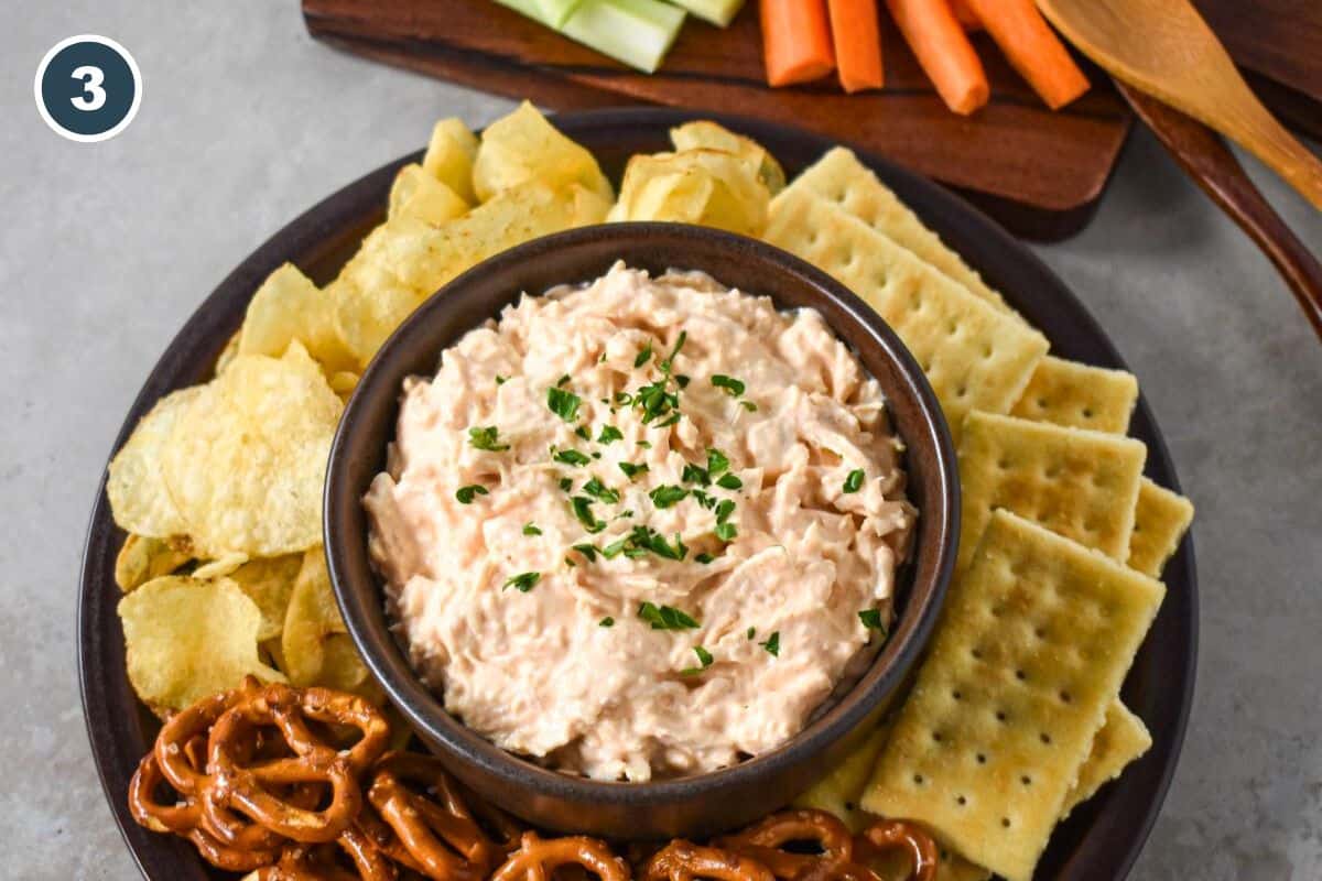 The dip served in a brown bowl served with pretzels, chips, crackers, carrots, and celery.