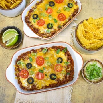 Layered taco dip in two small white baking dishes, topped with melted cheese, tomatoes, black olives, and green onions, served with tortilla chips and lime wedges on the side.