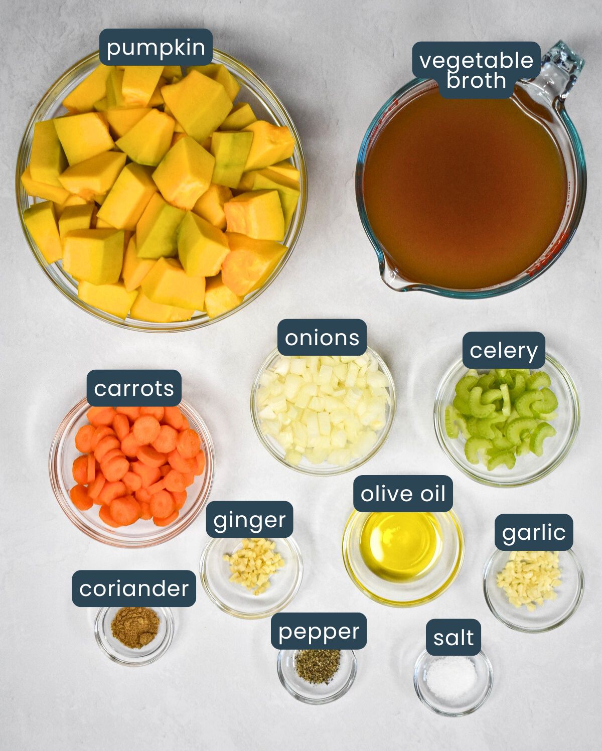 The ingredients for the soup prepped and arranged in glass bowls on a white table with each labeled with blue and white letters.