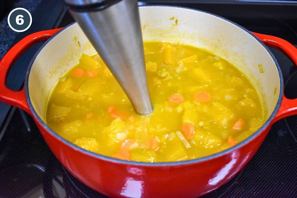 The pumpkin ginger soup being processed with an immersion blender in a red pot.