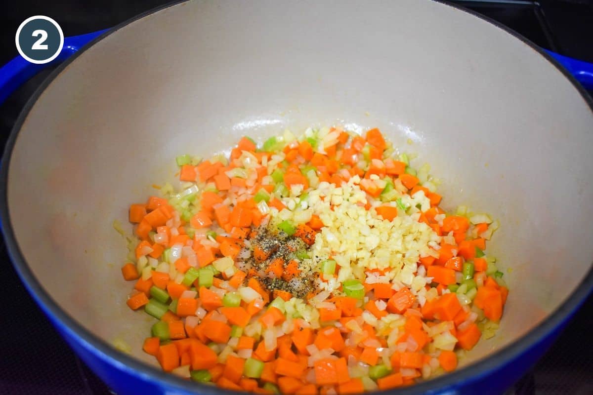 Minced garlic and black pepper added to the carrots, onions, and celery cooking in a large, blue and white pot.