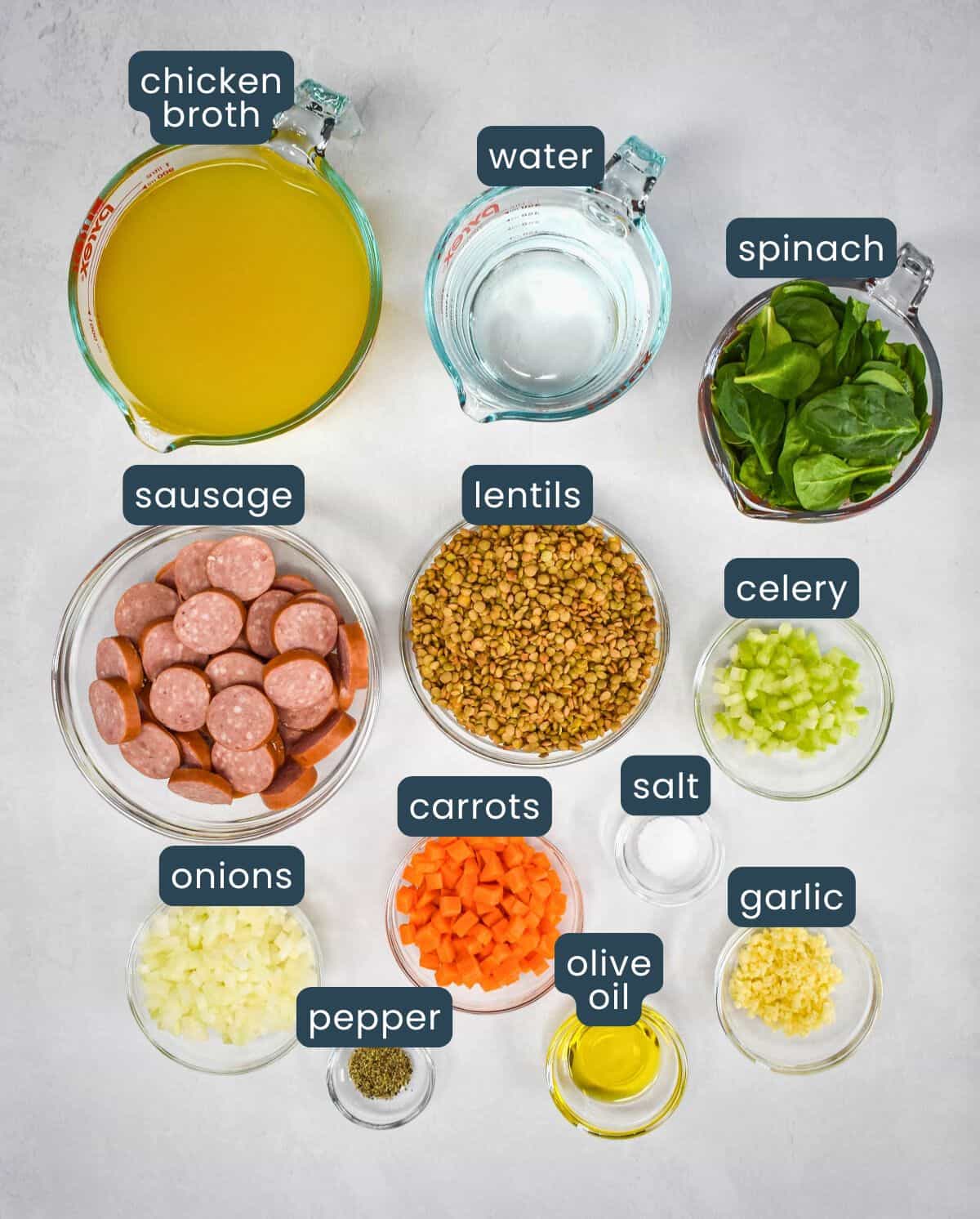 The prepped ingredients for the soup arranged on a white table in glass bowls with each labeled in blue and white letters. 