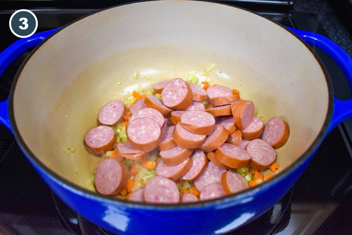 Sausage rounds added to the onions, carrots, and celery in a large blue pot.