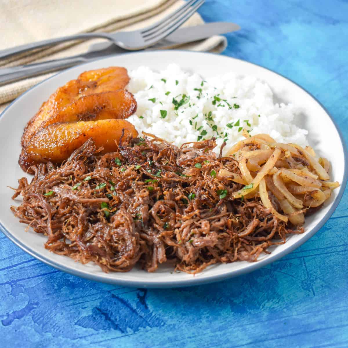 The vaca frita served with white rice, sauteed onions and fried sweet plantains on a white plate.