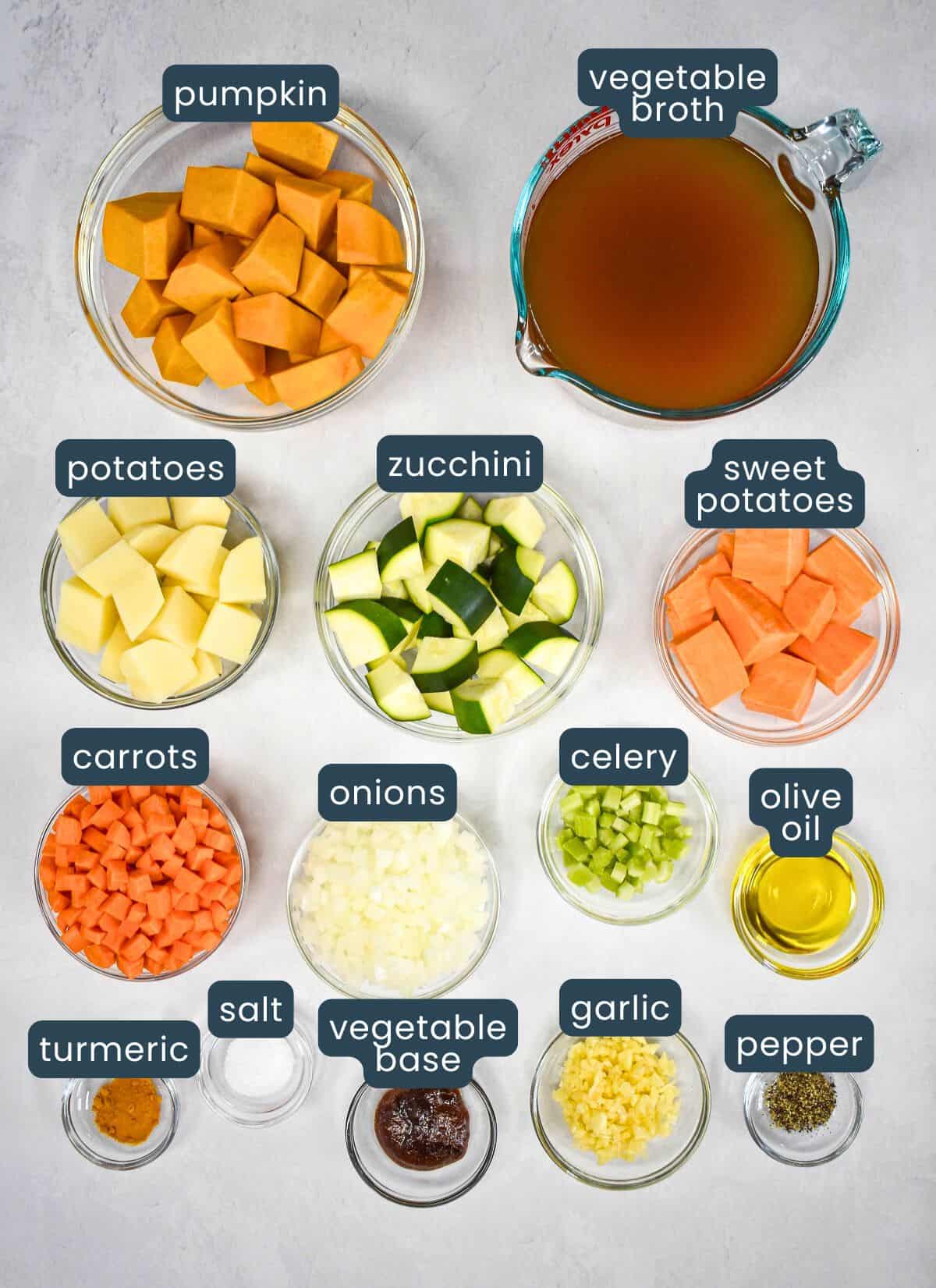 The prepped ingredients for the soup in glass bowls on a white table with each labeled with the name.