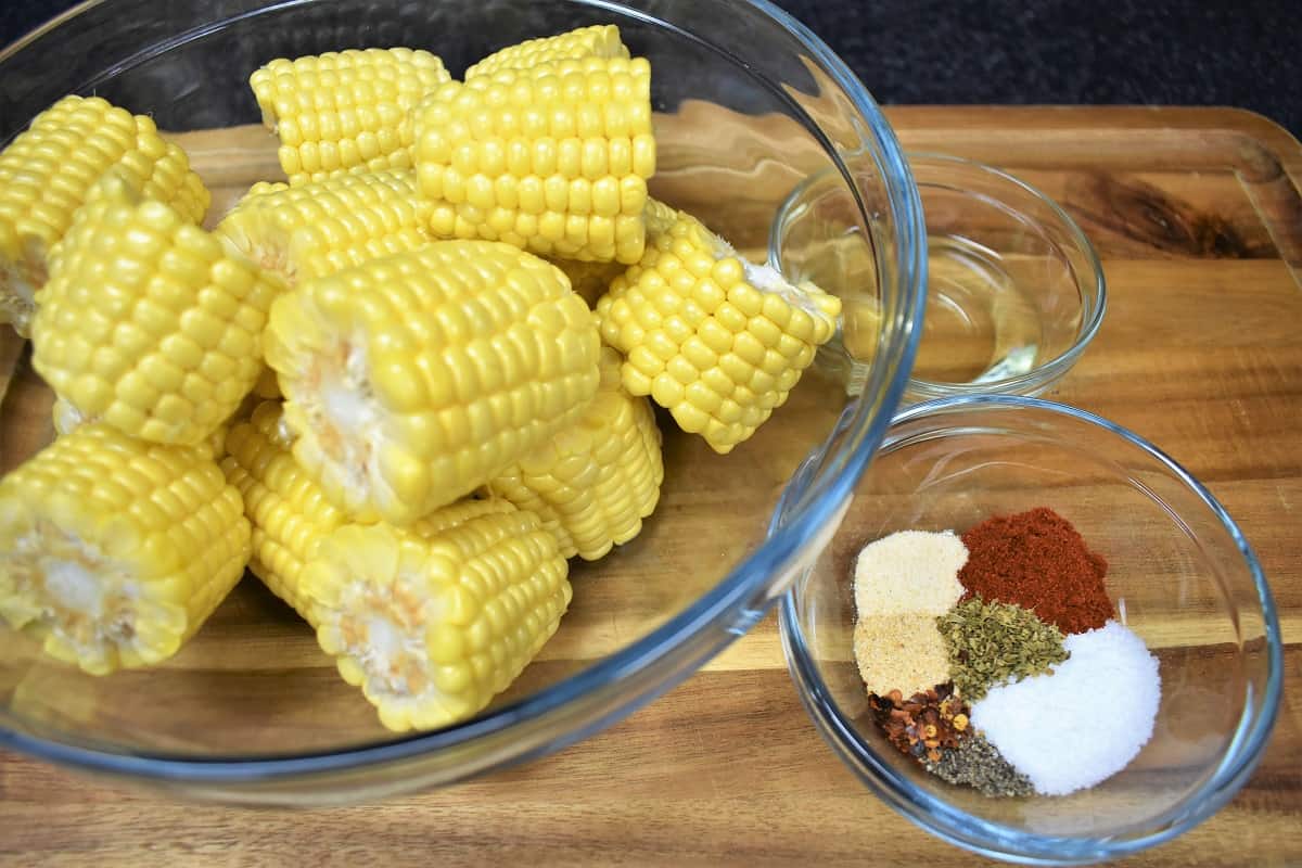 Cut corncobs in a glass bowl and a small bowl of seasoning displayed on a wood cutting board.