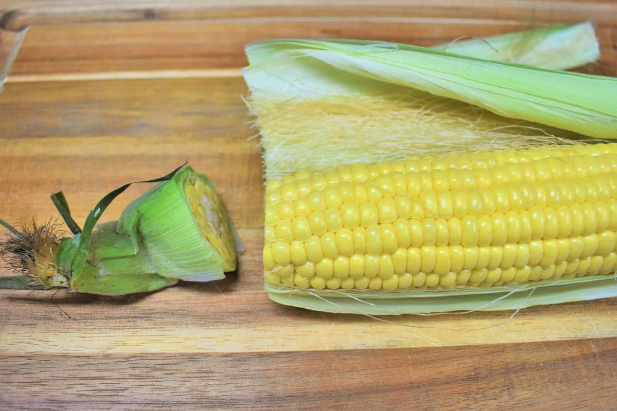 A fresh corn cob with half the husk removed, displayed on a wood cutting board.