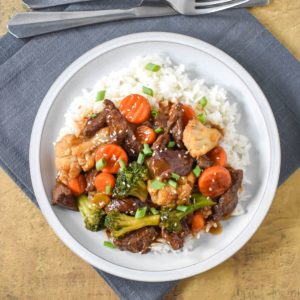 The beef and vegetable stir fry served over white rice on a white plate and garnished with sesame seeds and sliced green onions.