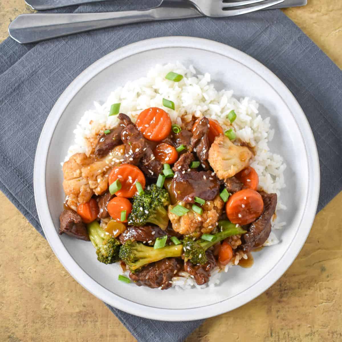 The beef and vegetable stir fry served over white rice on a white plate and garnished with sesame seeds and sliced green onions.