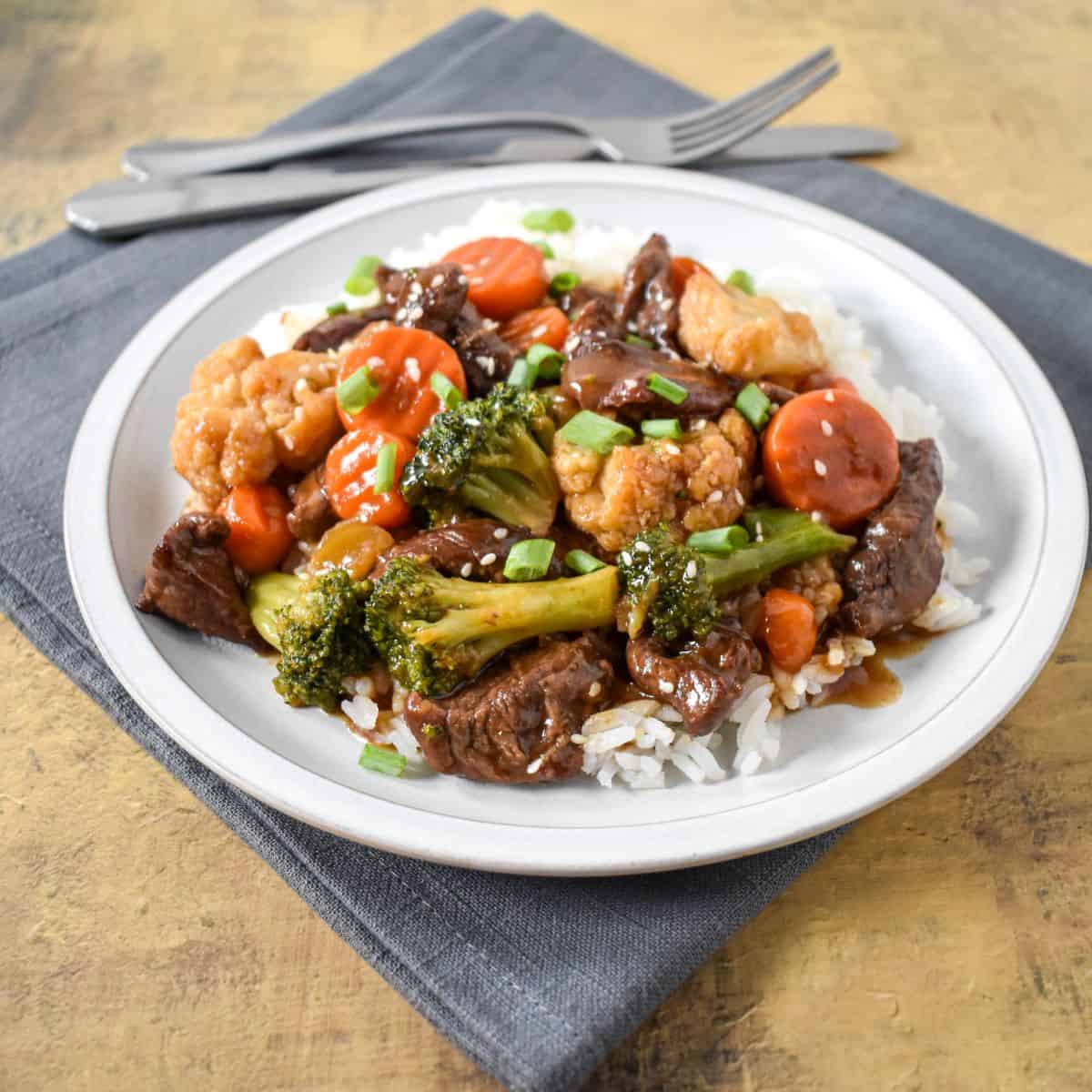 The finished beef stir fry served over white rice, garnished with sesame seeds and sliced onions, and served on a white plate.