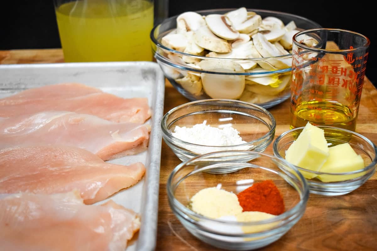 Thin chicken breasts, sliced mushrooms, spices, butter, flour, chicken broth and olive oil measured out and displayed on a wood cutting board.