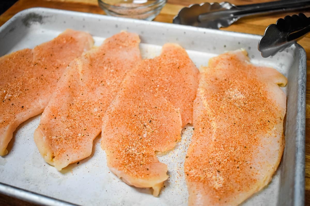 Seasoned, thin chicken breasts on a metal baking sheet.