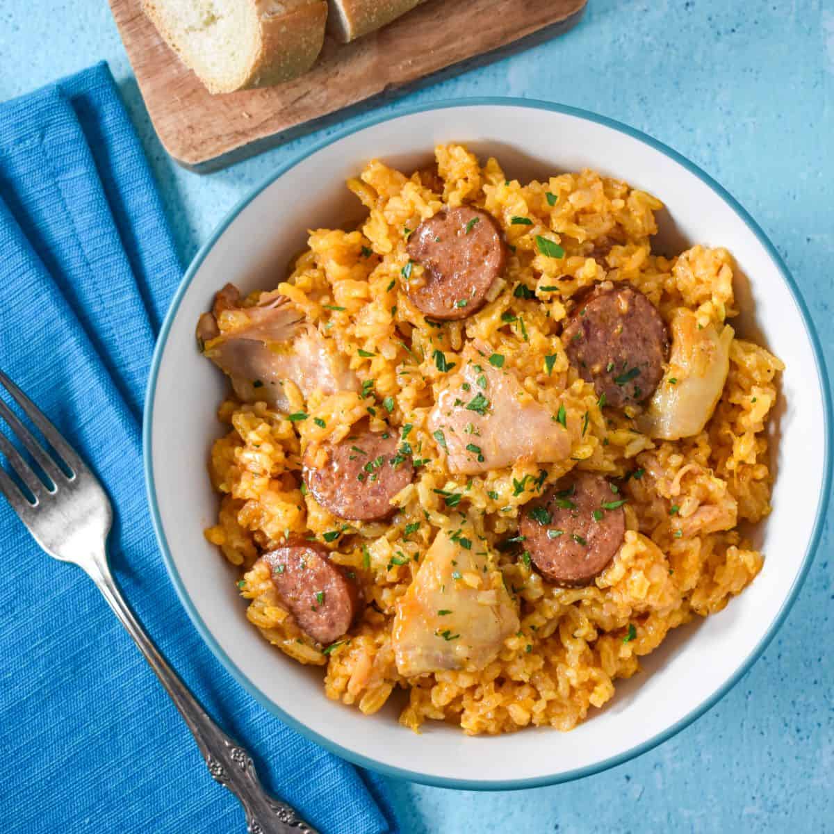 The chicken and rice with sausage served in a white bowl and set on a blue table with a teal linen to the left and bread.