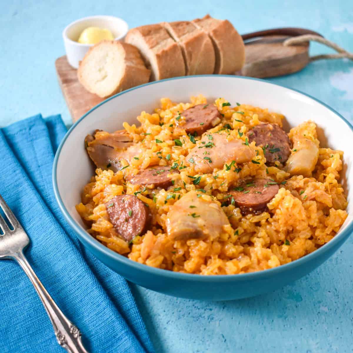 The chicken and rice with sausage served in a white bowl and set on a blue table with a teal linen to the left and bread.