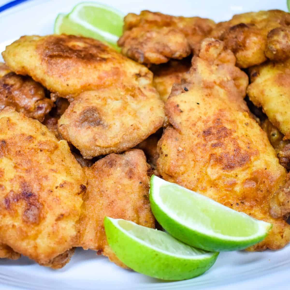 Fried chicken thighs stacked on a white platter with lime wedges for garnish.