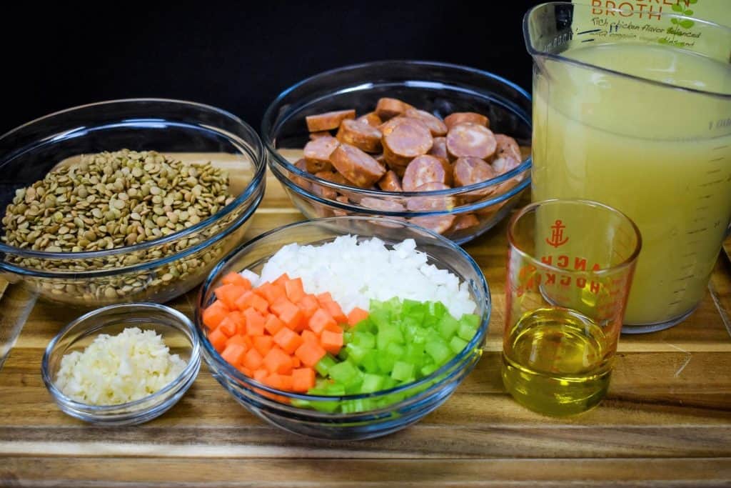 The ingredients for the lentil andouille sausage soup prior to cooking, arranged in clear glass bowls on a wood cutting board.