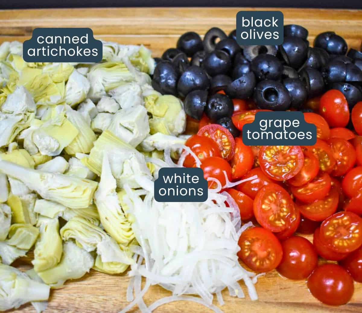 Quartered artichokes, grape tomatoes cut in half, black olives and thinly sliced onions displayed on a wood cutting board.