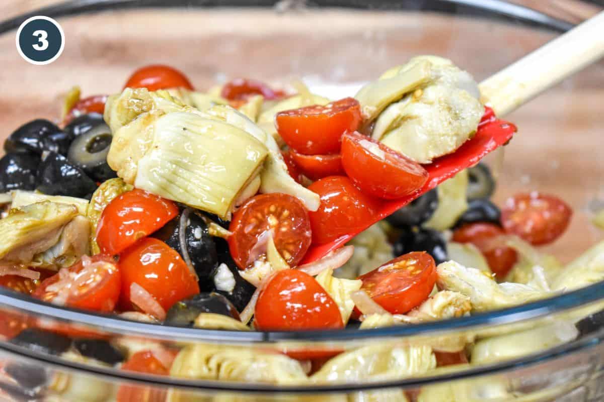 The artichoke salad being tossed with a red rubber spatula in a clear glass bowl.