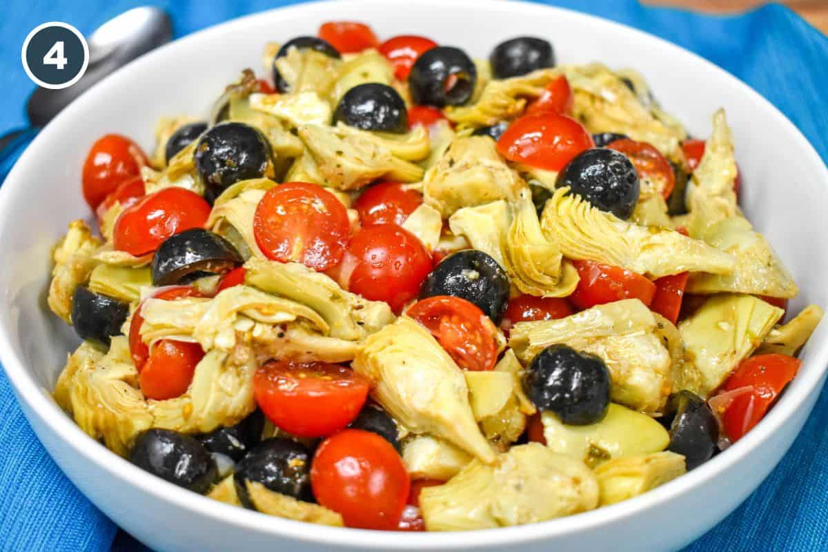 A cold salad with quartered artichokes, grape tomatoes, black olives and sliced onions tossed with an oil and vinegar dressing and served in a large white bowl on a teal linen.