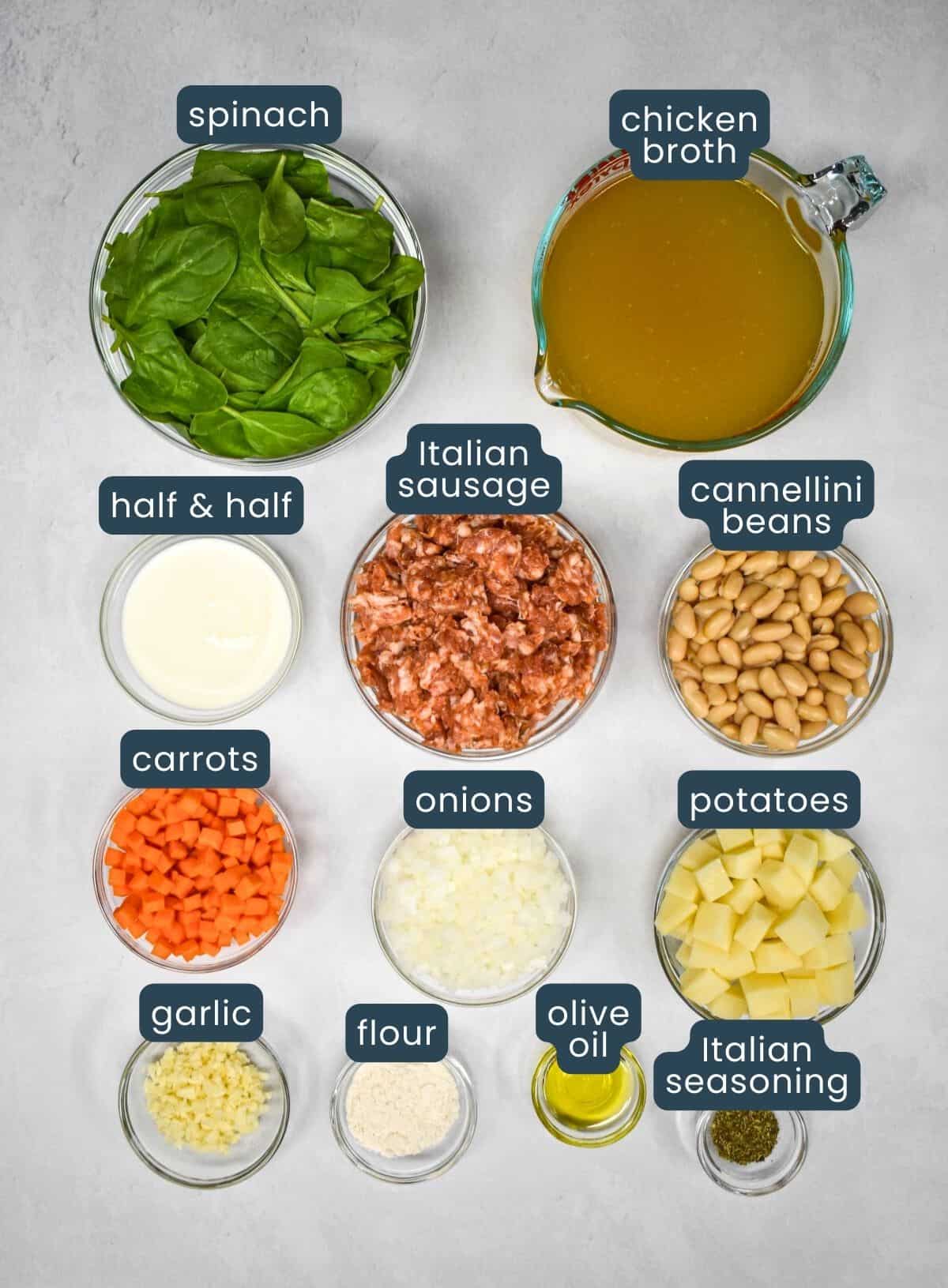 The ingredients for the tuscan soup prepped and arranged in glass bowls on a white table with each labeled with blue and white letters.