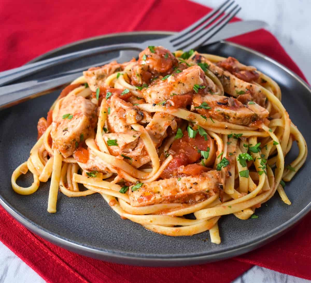 Linguine noodles tossed with tomato sauce, sliced chicken breasts and garnished with parsley, served on a gray plate.