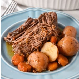 A piece of pot roast served with carrots and small red potatoes on a light blue plate.