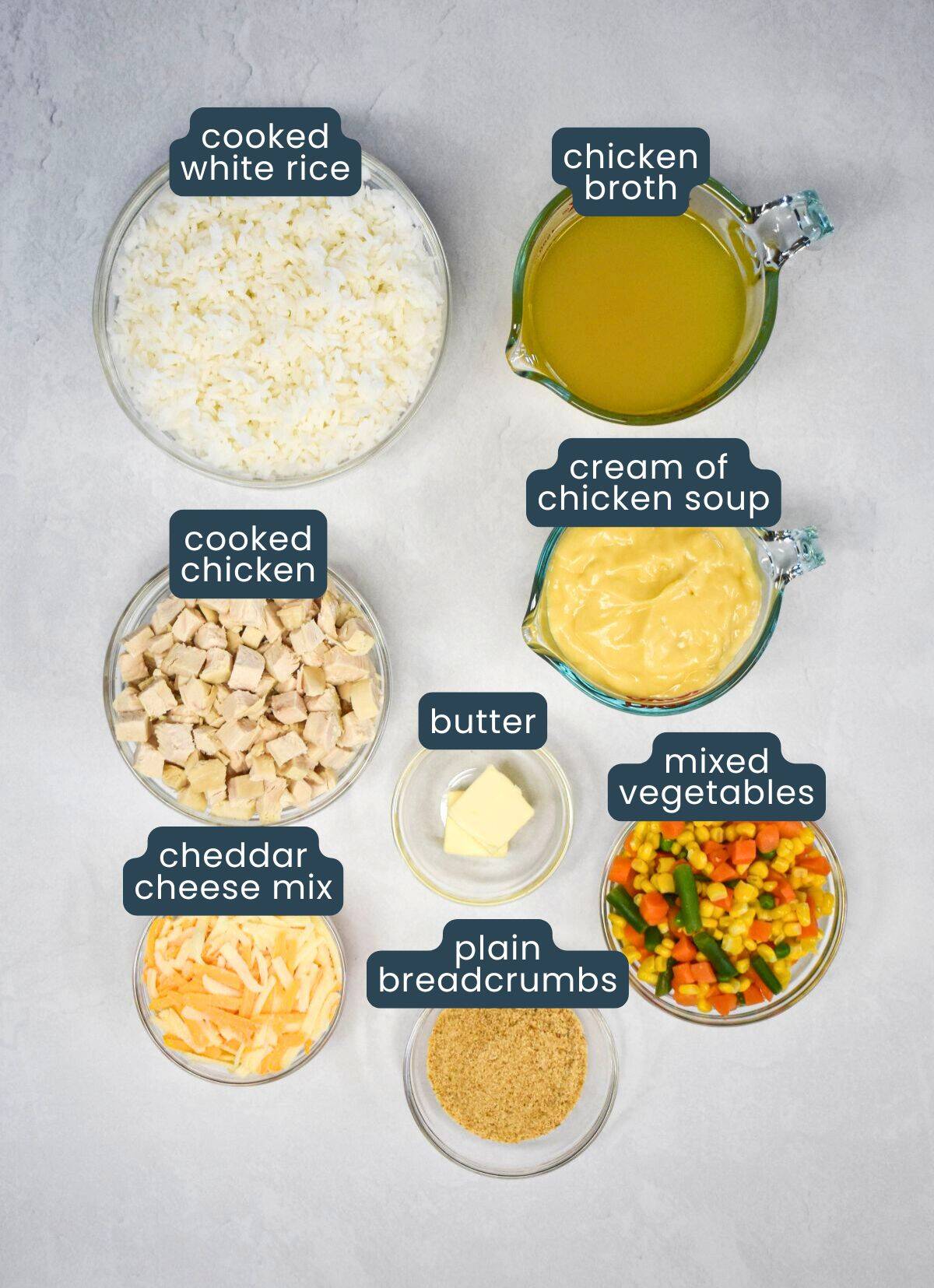 Labeled ingredients for creamy chicken and rice casserole, including cooked chicken, white rice, chicken broth, cream of chicken soup, cheddar cheese, butter, mixed vegetables, and breadcrumbs.