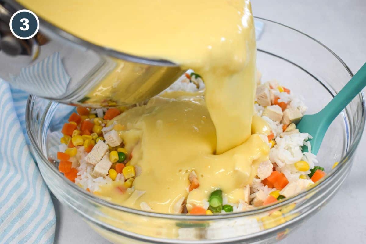 Pouring creamy sauce over chicken, rice, and mixed vegetables in a glass bowl.