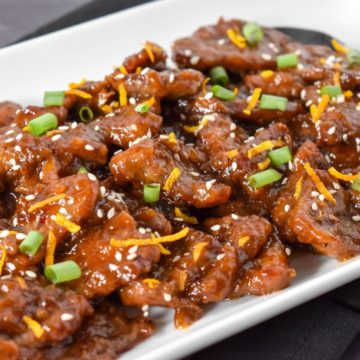 The orange beef garnished with orange zest, sesame seeds and sliced green onions and served in a rectangular, white platter.