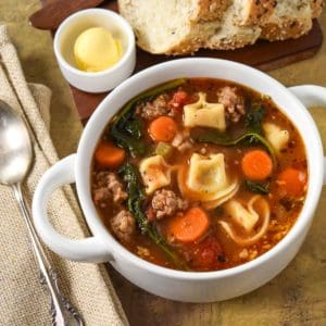 The sausage tortellini soup served in a white bowl served with sliced bread and butter.