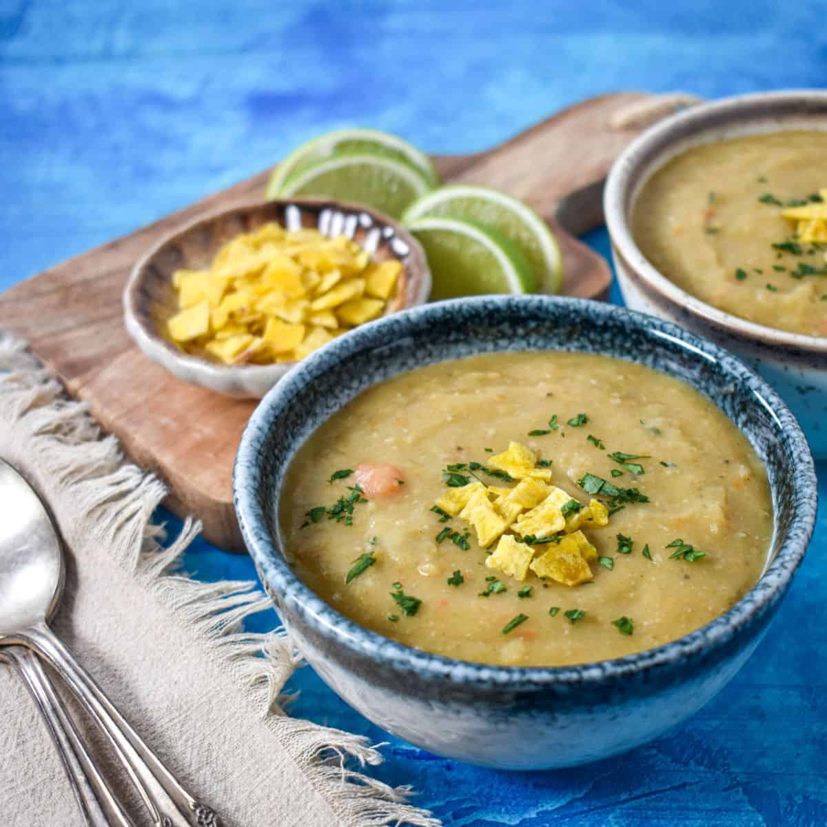 A bowl of creamy soup garnished with crushed plantain chips and fresh herbs, served with lime wedges on the side.