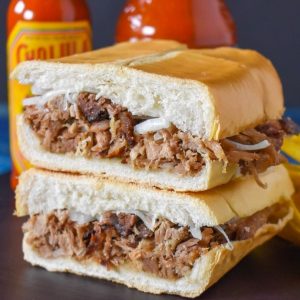 The roast pork sandwich cut in half and stacked with two bottles of hot sauce in the background.