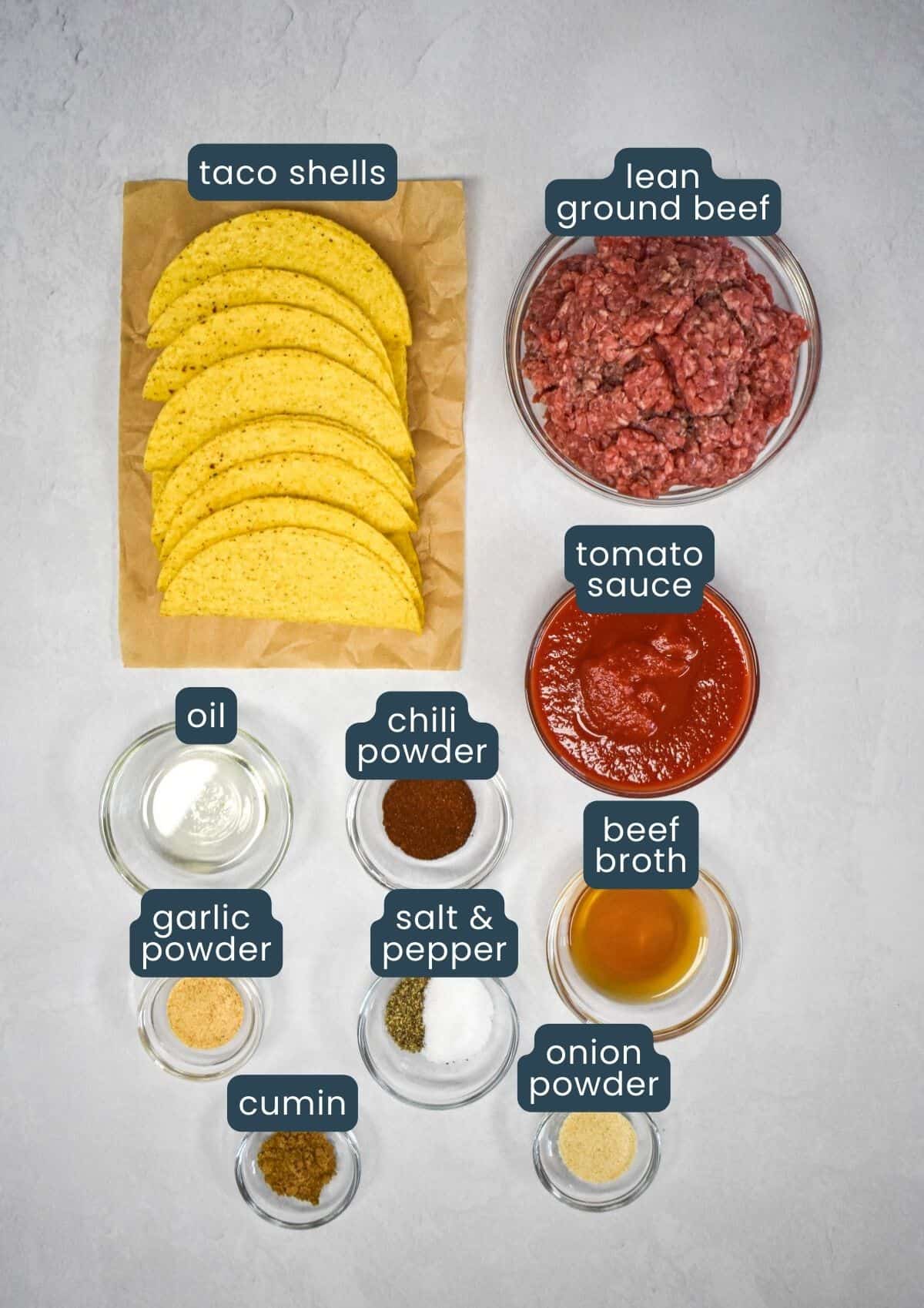 The ingredients for the tacos arranged in glass bowls and set on a white table with each one labeled with blue and white letters.