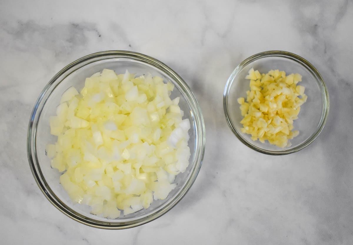 Diced onions and minced garlic displayed in glass bowls on a white table.