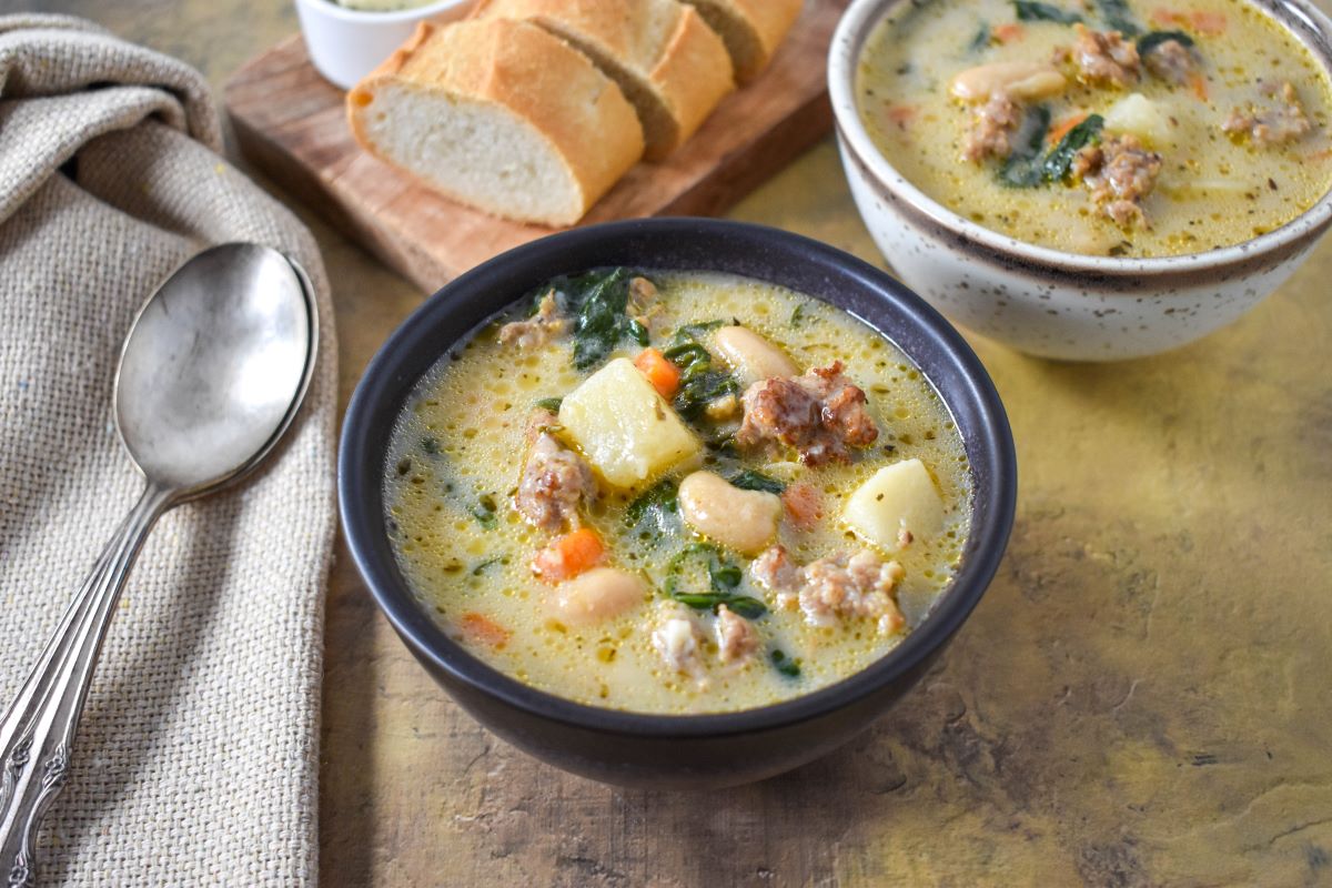 The finished Tuscan soup with cannellini beans served in a dark brown bowl with bread and another bowl of soup in the background.