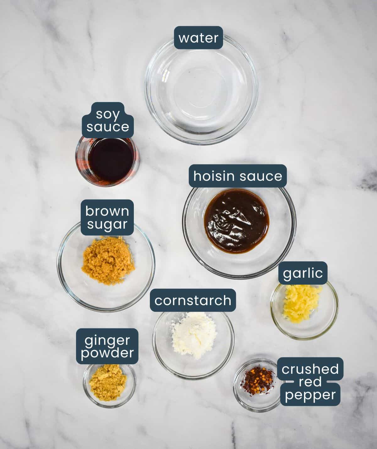 The ingredients for the stir fry sauce arranged in glass bowls on a white table with each labeled in blue and white.