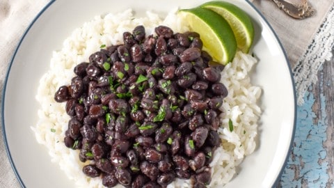 An image of black beans over white rice with two lime wedges, served on a white plate with a blue rim. The plate is set on a blue wood table with a beige linen and a fork to the top right.