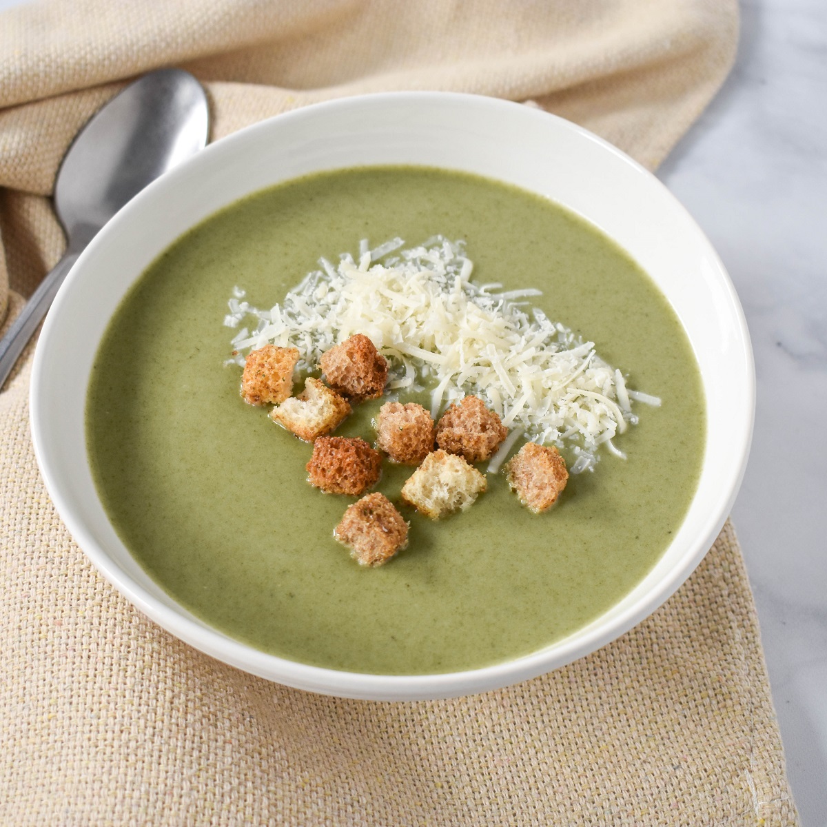 A bowl of cream of spinach soup topped with shredded Parmesan cheese and crispy croutons, served on a beige napkin with a spoon on the side.