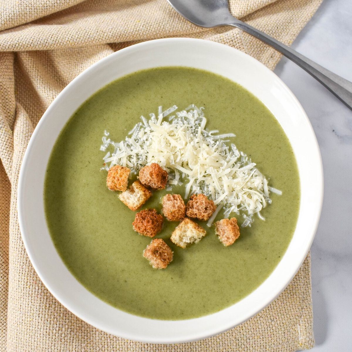 A bowl of cream of spinach soup, garnished with shredded Parmesan cheese and crispy croutons, served in a white dish with a spoon on the side.