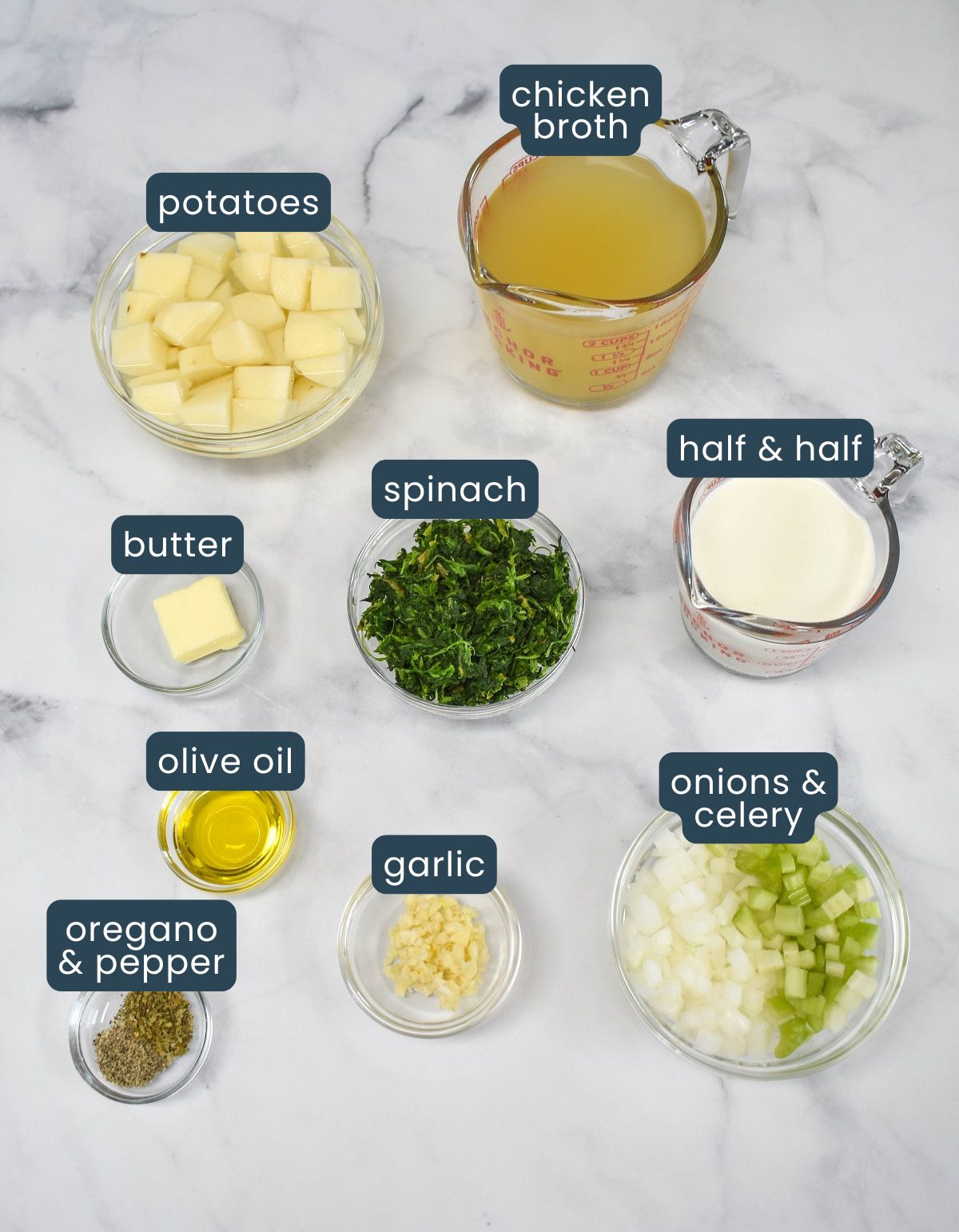 Labeled ingredients for cream of spinach soup arranged on a white marble surface, including potatoes, chicken broth, half & half, spinach, onions, celery, garlic, olive oil, butter, and seasonings.