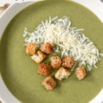 Cream of spinach soup in a white bowl, garnished with shredded Parmesan cheese and crispy croutons. Text overlay with the title on top.